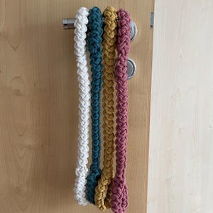 May include: Four long, braided cotton cords hanging vertically from a silver metal door handle. The cords are colored white, teal, mustard yellow, and dusty rose. Each cord features a thick, uniform, woven texture created by a repeating knot pattern.