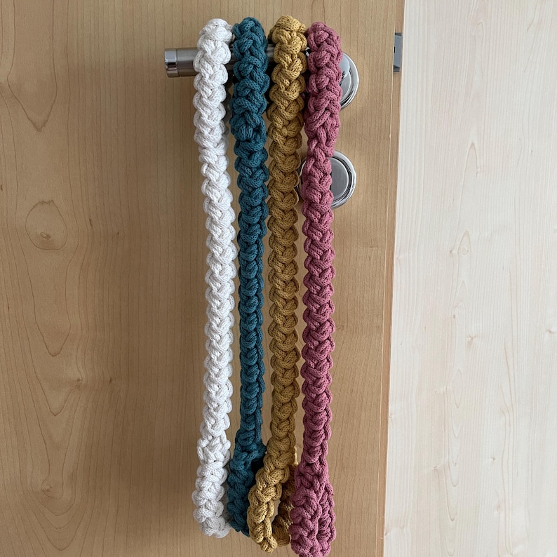 May include: Four long, braided cotton cords hanging vertically from a silver metal door handle. The cords are colored white, teal, mustard yellow, and dusty rose. Each cord features a thick, uniform, woven texture created by a repeating knot pattern.