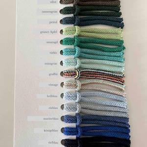 Puede incluir: Una carta de colores titulada Splendid Farbkarte que presenta una fila vertical de diecis&eacute;is muestras de cord&oacute;n trenzado en varios tonos de verde, azul y patrones multicolores. Cada cord&oacute;n est&aacute; anudado a una tarjeta blanca con etiquetas.
