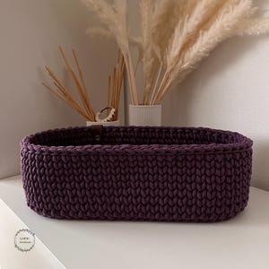 Puede incluir: Una cesta de almacenamiento de forma ovalada tejida a ganchillo con hilo grueso de color morado oscuro. La cesta presenta una textura de punto consistente en todas sus paredes exteriores e incluye un peque&ntilde;o detalle de leng&uuml;eta de cuero.