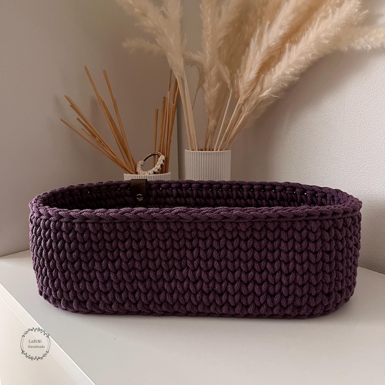 Puede incluir: Una cesta de almacenamiento de forma ovalada tejida a ganchillo con hilo grueso de color morado oscuro. La cesta presenta una textura de punto consistente en todas sus paredes exteriores e incluye un peque&ntilde;o detalle de leng&uuml;eta de cuero.
