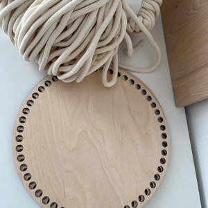 May include: A round wooden loom with holes around the edge, used for crocheting or macrame. The loom is made of light wood and has a natural finish. A thick white rope is lying on top of the loom.
