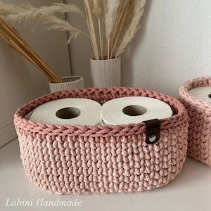 Puede incluir: Una cesta ovalada tejida a mano en un tono rosa suave, con un patr&oacute;n de punto grueso y texturizado. La cesta contiene dos rollos de papel higi&eacute;nico blancos e incluye una peque&ntilde;a etiqueta de cuero marr&oacute;n con un remache plateado. El texto dice Labini Handmade.