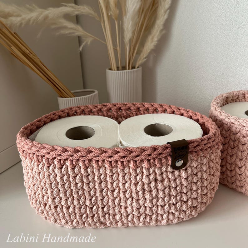 Puede incluir: Una cesta ovalada tejida a mano en un tono rosa suave, con un patr&oacute;n de punto grueso y texturizado. La cesta contiene dos rollos de papel higi&eacute;nico blancos e incluye una peque&ntilde;a etiqueta de cuero marr&oacute;n con un remache plateado. El texto dice Labini Handmade.