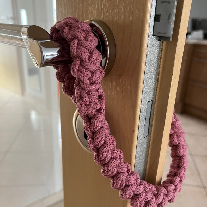May include: A dusty rose colored, thick, braided cord accessory looped around a silver metal door handle. The cord features a dense, repeating knot pattern throughout its length, creating a textured, rope-like appearance that hangs from the handle.
