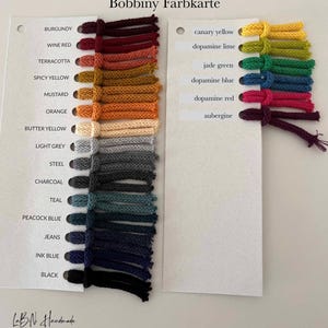 Puede incluir: Una carta de colores Bobbiny Farbkarte con dos tarjetas blancas y muestras de hilo adjuntas. La tarjeta izquierda enumera colores desde burdeos hasta negro, mientras que la derecha muestra colores desde amarillo canario hasta berenjena.