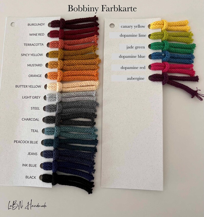 Puede incluir: Una carta de colores Bobbiny Farbkarte con dos tarjetas blancas y muestras de hilo adjuntas. La tarjeta izquierda enumera colores desde burdeos hasta negro, mientras que la derecha muestra colores desde amarillo canario hasta berenjena.