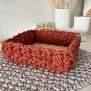 May include: A square, rust-colored crochet basket with a star-like stitch pattern and a flat wooden base. The basket features a small, dark brown leather-like tag on the side. It sits atop a larger, light gray crochet mat with a matching stitch.