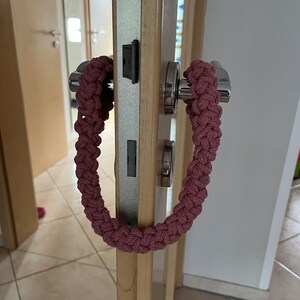 May include: A dusty rose-colored braided cord loop hanging from a silver metal door handle. The cord features a thick, intricate woven pattern made from a continuous length of rope, forming a large, flexible oval shape suspended from the lever.