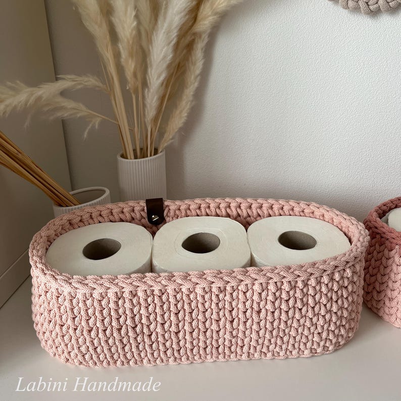 Puede incluir: Una cesta de almacenamiento ovalada de ganchillo rosa claro que contiene tres rollos de papel higi&eacute;nico blanco. La cesta presenta una leng&uuml;eta de cuero marr&oacute;n oscuro con un remache met&aacute;lico en el borde superior. El texto Labini Handmade es visible.