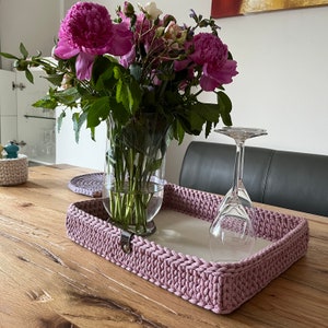 Könnte beinhalten: Ein rechteckiges, mauvefarbenes Häkeltablett mit hellem Innenraum steht auf einem Holztisch. Eine klare Glasvase enthält rosa Pfingstrosen und Grünzeug. Ein umgedrehtes Weinglas liegt auf dem Tablett.