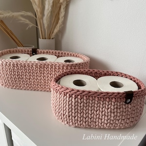 Puede incluir: Dos cestas ovaladas de ganchillo en color rosa empolvado con un patr&oacute;n de punto grueso y texturizado. Cada cesta presenta una leng&uuml;eta de cuero marr&oacute;n oscuro asegurada con un remache de metal plateado. Las cestas contienen rollos de papel higi&eacute;nico.