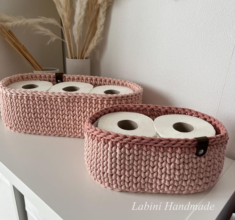 Puede incluir: Dos cestas ovaladas de ganchillo en color rosa empolvado con un patr&oacute;n de punto grueso y texturizado. Cada cesta presenta una leng&uuml;eta de cuero marr&oacute;n oscuro asegurada con un remache de metal plateado. Las cestas contienen rollos de papel higi&eacute;nico.
