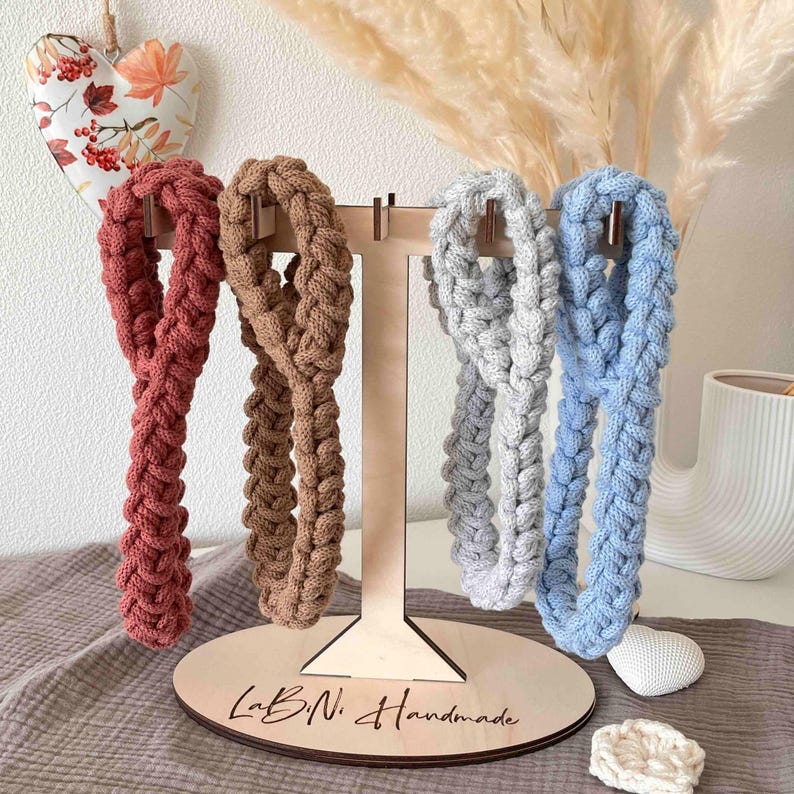 May include: Four braided headbands in rust, tan, light gray, and light blue hanging on a wooden T-shaped display stand. The base of the stand is oval-shaped and features the black text "LaBiNi Handmade" printed on the surface.