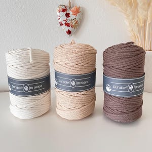 Könnte beinhalten: Drei Spulen geflochtenes Makramee-Garn in Creme, Beige und Braun. Jede Spule ist mit einem grauen Etikett mit der Aufschrift "Durable Braided" versehen. Die Kordeln sind eng gewickelt und zeigen ihre Textur. Im Hintergrund hängt ein dekoratives Herz mit floralen Akzenten.
