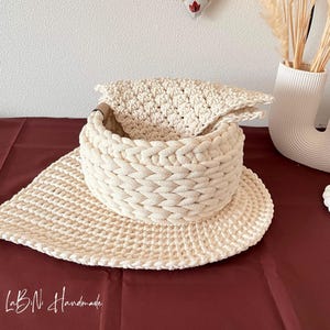 May include: A cream-colored crocheted basket with a thick braided rim and a flat, leaf-shaped base. The basket features a textured, woven pattern and includes a matching crocheted cloth insert. The text LaBiNi Handmade is visible in the corner.