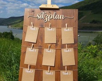 May include: A wooden easel with a brown wooden sign that says "Sitzplan" in white lettering. The sign has nine brown paper cards with white text that says "Tisch 1" through "Tisch 9" hanging from string with clothespins.