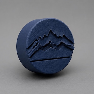 May include: A navy blue, cylindrical bottle stopper with a mountain range design carved into the top. The stopper is made of a solid material and has a textured surface. The mountain range design features three layers of peaks.