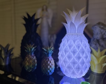 May include: A collection of pineapple-shaped lamps, including a large white lamp with a geometric pattern, a smaller iridescent lamp, and a yellow lamp. The lamps are on a black surface with a reflection of the white lamp.