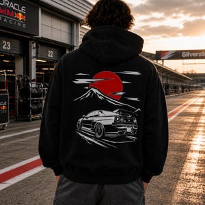 Könnte beinhalten: Schwarzer Kapuzenpullover mit japanisch inspiriertem Design auf dem Rücken. Die Grafik zeigt eine rote Sonne, einen Berg und eine weiße Illustration eines Sportwagens. Der Hoodie wird in einer Rennumgebung getragen.