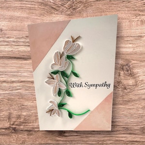 May include: A handmade sympathy card featuring quilled flowers in shades of cream and brown, with green stems and leaves. The card has a light pink and cream background with the words "With Sympathy" in black script.