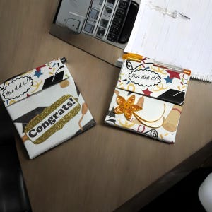 Puede incluir: Dos pequeños cuadernos blancos con diseños coloridos de temática de graduación. Un cuaderno tiene una pegatina de purpurina dorada que dice "¡Enhorabuena!" y el otro tiene una pegatina que dice "¡Lo hiciste!!".