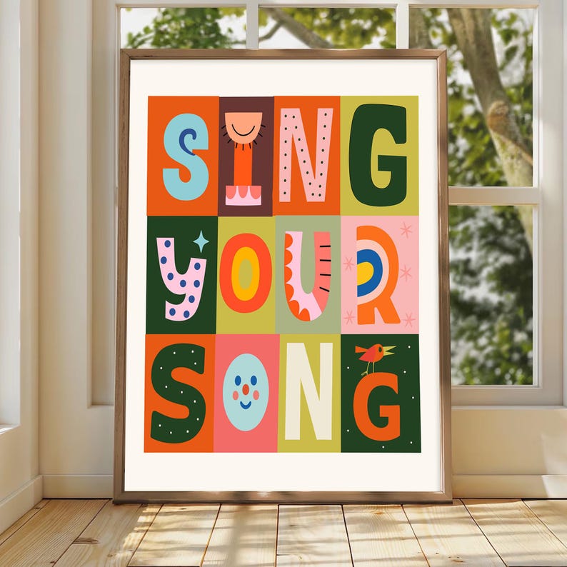May include: A framed art print with the words "SING YOUR SONG" in colorful, block letters. The letters are in various colors, including orange, green, blue, and pink, set against a grid of different colored squares. A small bird is perched on the letter "G".