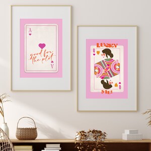 Op de afbeelding: Twee ingelijste prints met roze achtergronden en gouden lijsten. De linker print toont een speelkaart met een roze hart en de tekst "good for the plot". De rechter print toont een speelkaart met een hartenkoningin en de tekst "LUCKY ME".