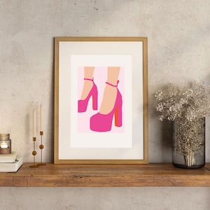 May include: A framed print of a pair of pink high-heeled shoes with ankle straps on a pink background.