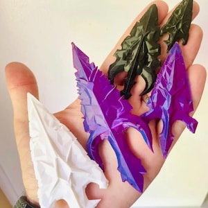 May include: A hand holds several decorative arrowheads in various colors. The arrowheads are white, black, and purple. The arrowheads have a detailed, textured design.