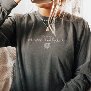 Pode incluir: Uma mulher vestindo uma camisa de manga comprida cinza escura com o texto "in my behavior analyst era" e um pequeno gráfico de um globo. Ela está sentada em um sofá e segurando uma caneca preta.