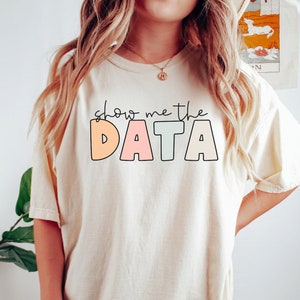 May include: A woman wearing a light beige t-shirt with the text "Show me the DATA" in colorful letters. The shirt is a relaxed fit and has a crew neck. The model is wearing the shirt with a pair of dark green leggings. The background is a white wall with a plant in a white pot.