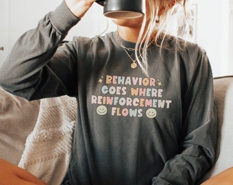May include: A dark grey long-sleeved shirt with the text "BEHAVIOR GOES WHERE REINFORCEMENT FLOWS" in colourful letters with smiley faces on either side of the text.