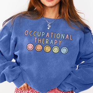 Könnte beinhalten: Ein blaues Sweatshirt mit dem Text "Occupational Therapy" und fünf bunten Smileys. Das Sweatshirt wird von einer Person getragen, die eine rot-weiß karierte Shorts trägt.