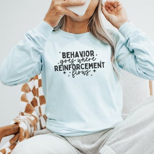 May include: A light blue long-sleeved shirt with the text "BEHAVIOR goes where REINFORCEMENT flows" printed on the front.