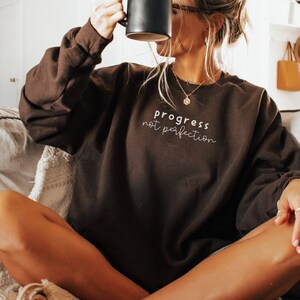 May include: A woman wearing a brown sweatshirt with the text "progress not perfection" in white lettering. She is sitting on a couch and holding a black mug.