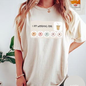 May include: A white t-shirt with the text "I am working for:" and a cartoon image of a coffee cup with a straw. There are five smiley faces below the text, each with a different color.
