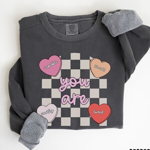 May include: A dark gray sweatshirt with the word "you are" in pink letters, surrounded by four heart-shaped candies with the words "capable", "smart", "worthy", and "loved". The background is a black and white checkered pattern. The sweatshirt has gray fleece cuffs.