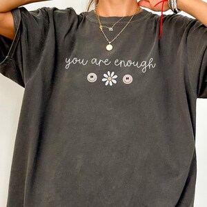 May include: An oversized t-shirt in a dark grey colour with the text "you are enough" and three smiley faces and a flower graphic.