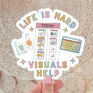 May include: A colorful sticker with the text "LIFE IS HARD VISUALS HELP". The sticker features a daily schedule, a thumbs-up graphic, and a tablet with symbols. The background is a woven mat.