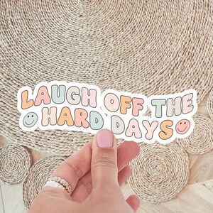 Könnte beinhalten: Ein bunter Aufkleber mit dem Text "Laugh off the hard days" mit Smileys auf beiden Seiten. Der Aufkleber wird von einer Hand vor einem gewebten Hintergrund gehalten.