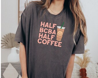 Puede incluir: Una camiseta gris oscuro con un gráfico rosa que dice "HALF BCBA HALF COFFEE" y una imagen de dibujos animados de una taza de café con una cara sonriente.