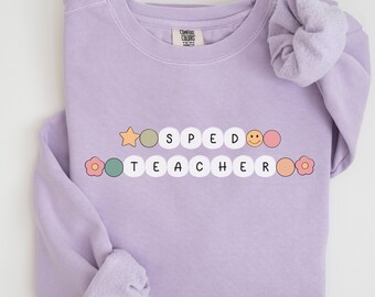 Könnte beinhalten: Fliederfarbenes Sweatshirt mit dem Text "SPED TEACHER" in einer verspielten, farbenfrohen Schriftart. Das Design umfasst einen Stern, ein Smiley und Blumen. Das Sweatshirt ist aus weichem Material und hat eine bequeme Passform.