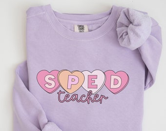 May include: A lavender sweatshirt with the words "SPED teacher" printed on it in pink and white letters. The letters are inside pink, peach, and white heart shapes.