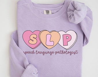May include: A lavender sweatshirt with a graphic of three pink, orange, and white candy hearts that spell "SLP" and the text "speech language pathologist".