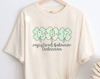 May include: A cream-coloured t-shirt with a graphic of three green shamrocks with the letters RBT inside. The text "registered behaviour technician" is written below the shamrocks.