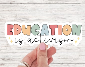 Può includere: Un adesivo bianco con il testo "EDUCATION is activism" in lettere colorate. Le lettere sono contornate di nero e hanno un leggero effetto ombra. Ci sono due piccole stelle dorate su entrambi i lati del testo.