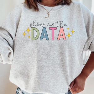 Puede incluir: Sudadera gris con el texto "show me the DATA" en colores arcoíris con estrellas amarillas a cada lado.