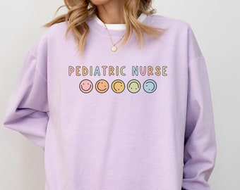 Puede incluir: Una sudadera de color lavanda con el texto "PEDIATRIC NURSE" en colores arcoíris y una fila de cinco caritas sonrientes arcoíris debajo del texto.