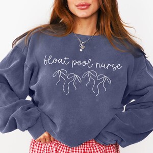 Könnte beinhalten: Ein marineblauer Pullover mit dem Text "float pool nurse" und drei Palmenillustrationen in Weiß. Der Pullover wird über einer roten und weißen karierten Shorts getragen.